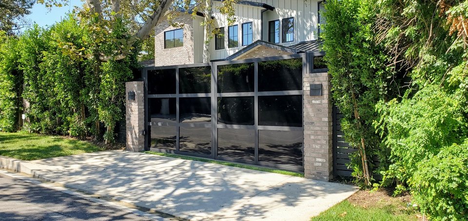 Black-framed driveway gate between pale masonry pillars and green hedges.